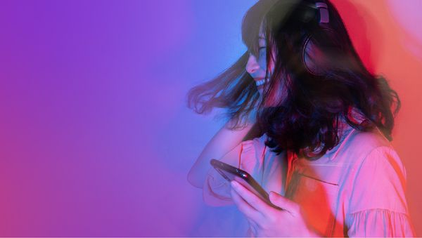 A young woman with music headphones on, holding a phone is dancing to music smiling with her eyes closed